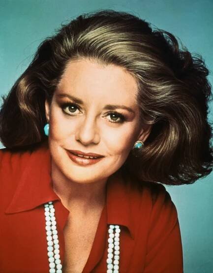 Portrait of Barbara Walters, in a red shirt with a white beaded necklace and blue earrings.