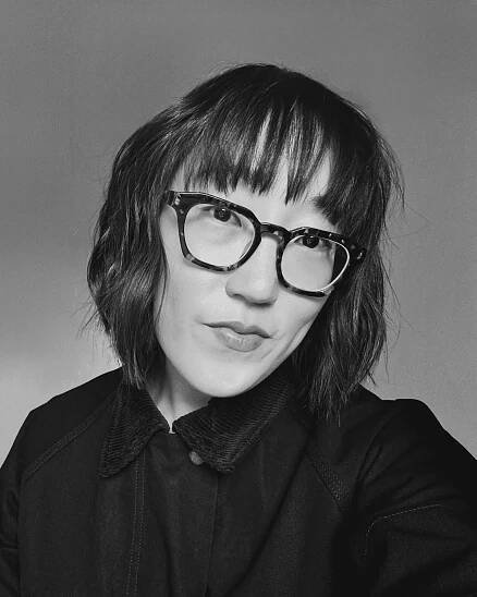 Sojin Rank, a Korean woman with short brown hair and bangs wearing large-framed glasses and a black jacket.