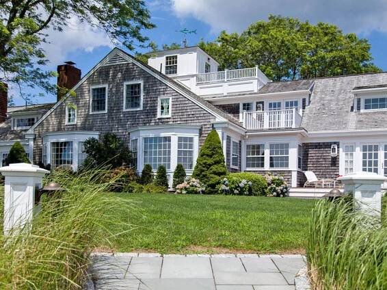 A two-story house with gray wooden shingles, white trim, and multiple windows. The house is surrounded by green lawn, shrubs, and trees under a partly cloudy sky. Two white pillars frame the entrance walkway.