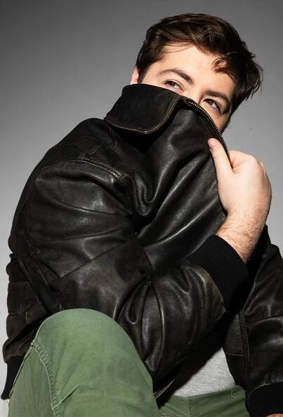 Michael Gandolfini covering the lower part of his face with a brown leather jacket. He's also wearing army green pants.