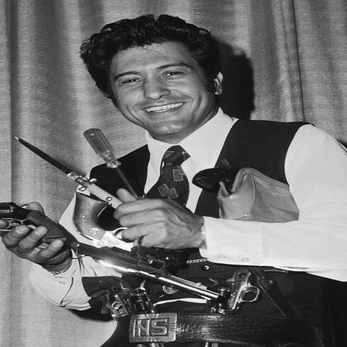 Nelson Sardelli in a vintage suit with a tie, holding props, smiling in front of a curtain backdrop