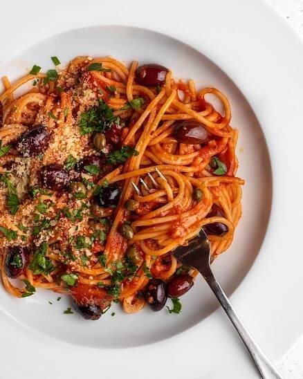 White plate with spaghetti in a puttanesca sauce. Fork with twirled noodles around it and fresh parsley and parmesan on top.
