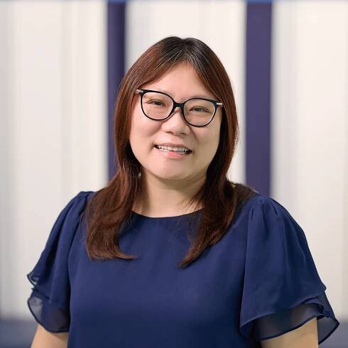 Lead Principal Ying Chen, wearing a dark blue blouse and glasses, standing in front of a dark blue striped white wall.