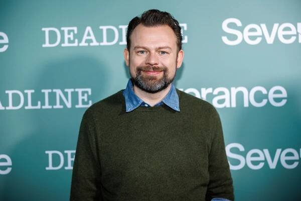 Dan Erickson at a media event wearing a sweater over a collared shirt, standing in front of a backdrop with "Deadline" and "Severance" text