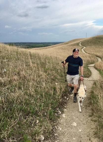 Paul is walking up a rocky trail surrounds by grass and hay. There is water far in the distance of the photo. He is holding his dog's leash in one hand and walking stick in the other hand. 