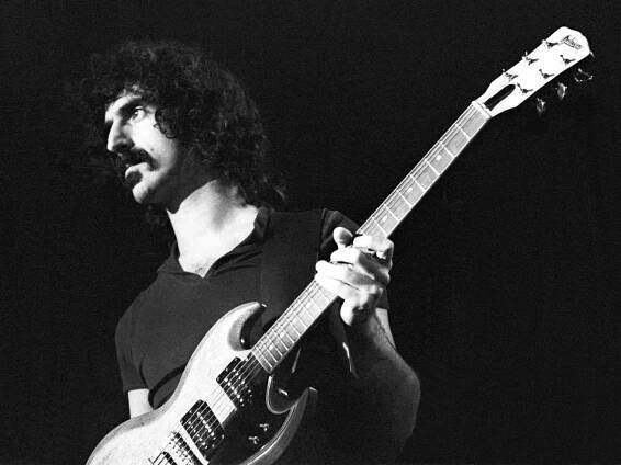 Archival photo of Frank Zappa. He is pictured in black and white, and is holding a Gibson SG.