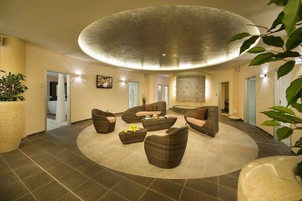 Wellness Hotel Diamant Hluboká nad Vltavou Exterior photo The photo shows a stylish and inviting hotel lobby or lounge area. The space is characterized by a circular seating arrangement in the center, surrounded by dark gray tiled flooring. The seating consists of several woven wicker armchairs and a sofa, 