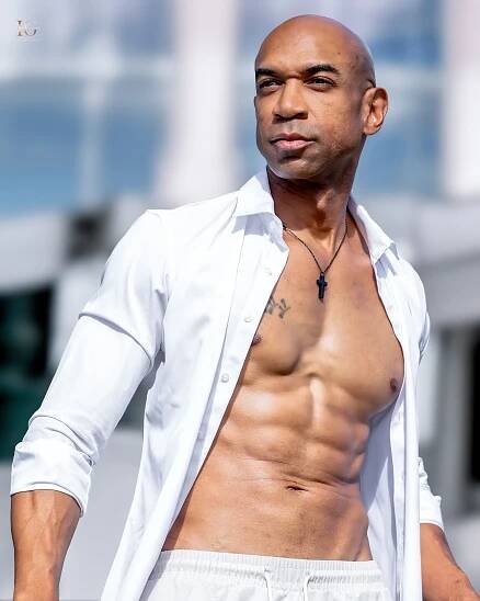 Athletic man in unbuttoned white shirt stands against blue background showing muscular physique in fashion pose.