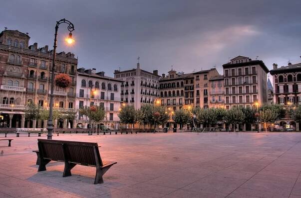 an empty bench sits in the middle of a plaza at dusk, with tall buildings behind it