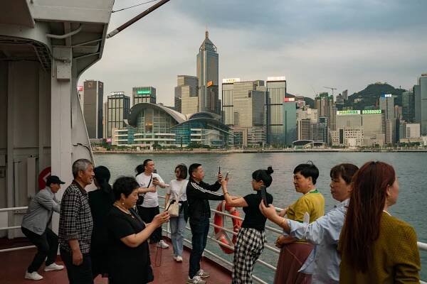 A group of tourists on a ferry in Hong Kong, several using their phones to take selfies with the city in the background.