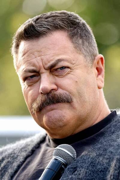 Person with a mustache holding a microphone, wearing a textured sweater, appears thoughtful against an outdoor backdrop