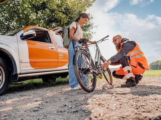 „Pannenhelfer in Warnweste repariert ein Fahrrad, während die Besitzerin daneben steht; im Hintergrund ein ACV-Servicefahrzeug.“