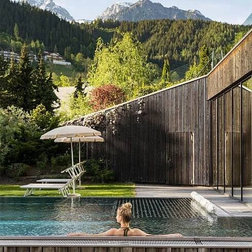 Frau entspannt sich im Schwimmbad mit Bergblick, umgeben von Holzbarrieren und Sonnenschirmen.