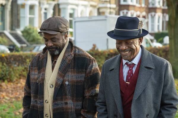 Two very well-dressed older men wearing hats smile and walk side-by-side through a city park. 