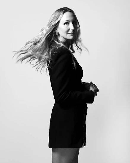 In a full-length black-and-white portrait, a woman in a short black outfit appears to be midway through turning toward the camera. Her hair is midair and she has a neutral expression.