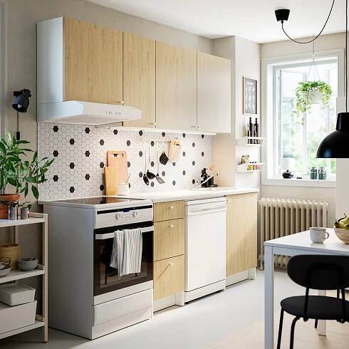 Moderní kuchyňská linka se dřevem, bílé spotřebiče, černé akcenty. Nerezová trouba, závěsné světlo, rostlina. Černý tečkovaný backsplash, bílý pult.