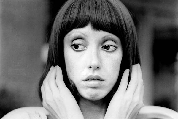 Shelley Duvall in a black-and-white portrait from the 1970s, holding her hands up to the sides of her head.