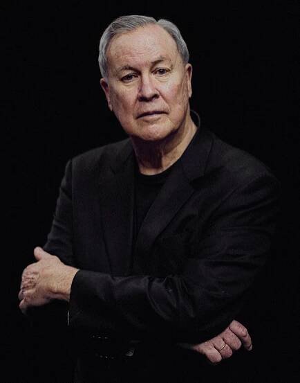Robert Wilson with arms folded wearing a black shirt and jacket on a black background.