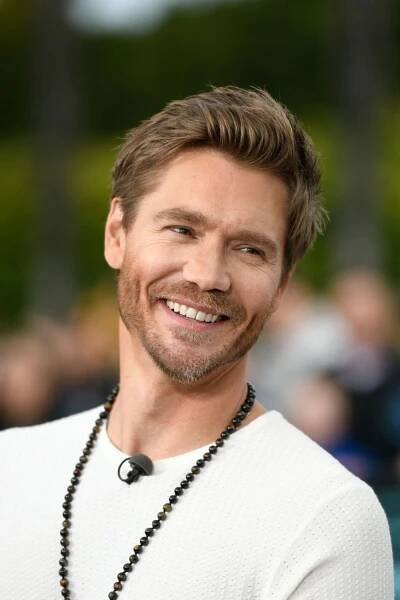a man with a smile on his face wearing a white sweater and black beaded necklace
