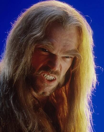 Person with long hair and a fierce expression, lips curled in a growl, against a plain background. Likely a dramatic or fantasy character