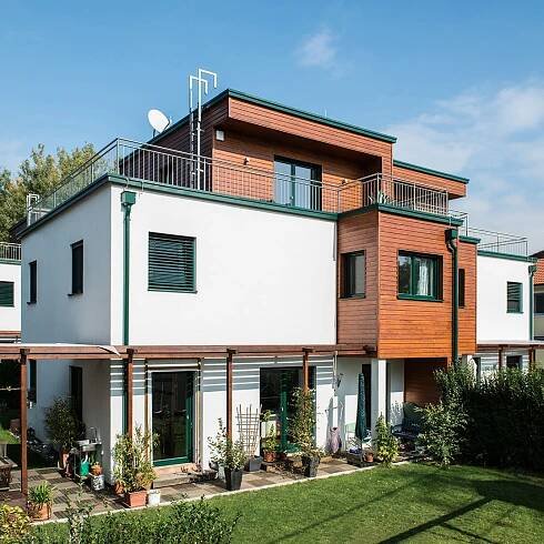 Moderne WOLF Haus mit nachhaltiger Bauweise, zweistöckiges Gebäude mit weißer Fassade und hölzernen Akzenten, begrünter Garten mit Terrassenbereich.