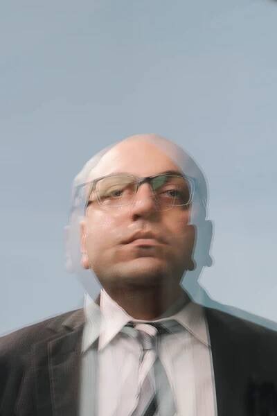 A double exposure portrait of a man with glasses in a suit against a blue-gray sky, seen from the chest upward. The man is in his 30s, bald, and wears a dark suit with a tie.