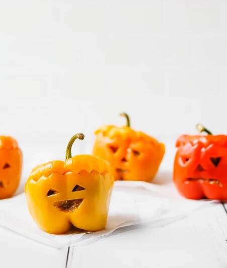 Spooky vegan bell pepper jack o'lanterns smiling at the camera on a white wooden table.