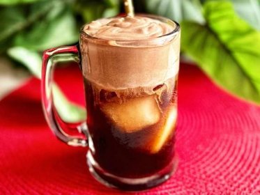 closeup of iced coffee in clear coffee mug with thick creamy chocolate foam on red placemat with greenery in background