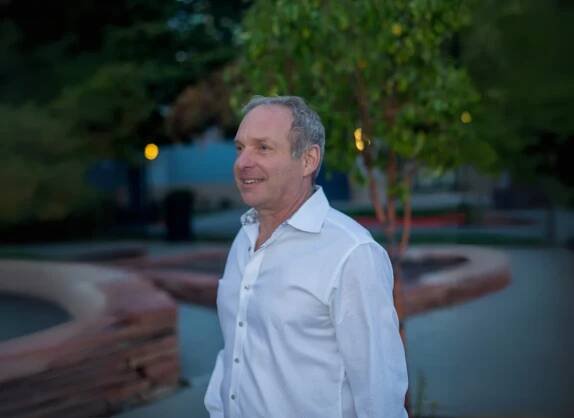 Eric Martin smiling outdoors in a white shirt, standing in a serene park setting with greenery and soft evening light.