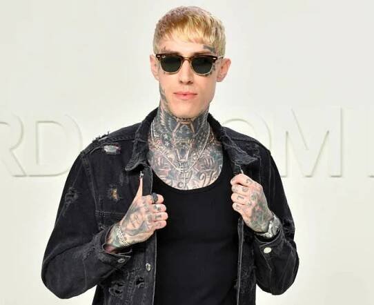 Person with tattoos and sunglasses, wearing a distressed denim jacket and jeans, posing confidently in front of a neutral background