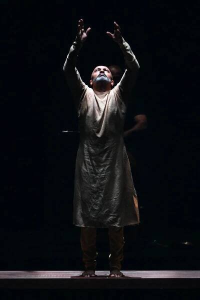 A dancer in a tunic raises his arms and looks up. The background is dark.