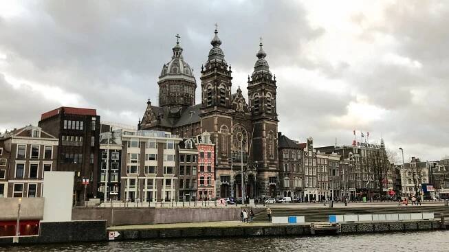 Cityscape of Basilica of Saint Nicholas, a historic building with tall spires and intricate architecture near a river, under a cloudy sky.