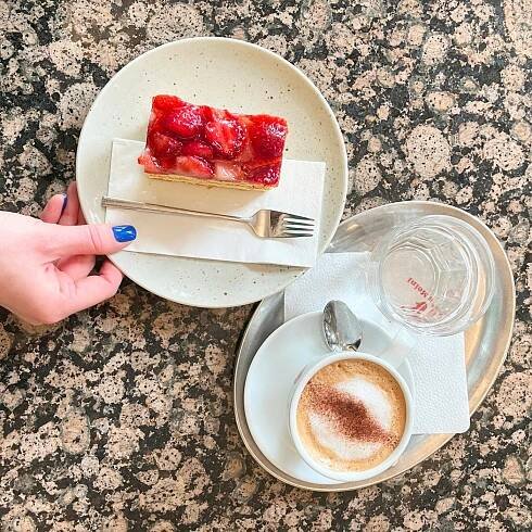 Ein Stück Erdbeerkuchen auf einem Teller und ein Cappuccino mit einem Glas Wasser auf einem Tablett, serviert auf einem Cafétisch.: 