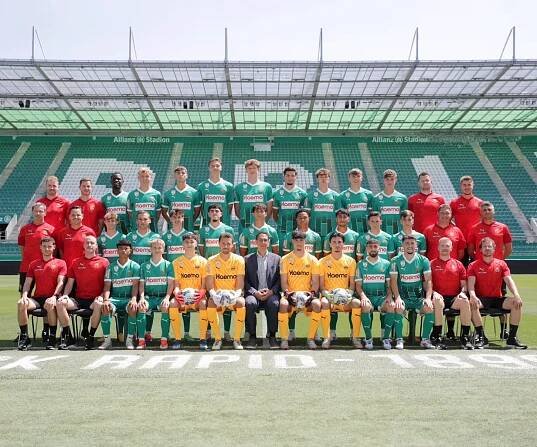 Fußballmannschaft in grünen Trikots posiert für ein Mannschaftsfoto mit Sponsor Mag. Markus Mateyka von der Firma Haemo Pharma im Allianz Stadion, begleitet von Trainern und Betreuern in roten Shirts.