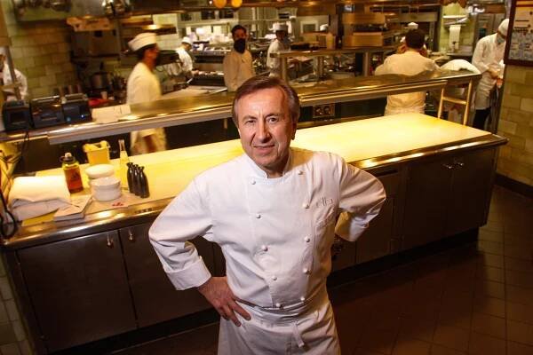 Daniel Boulud stands in a kitchen wearing a chef's jacket, hands on hips. 
