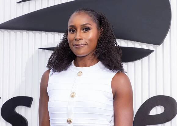 Person in a sleeveless, buttoned dress poses on a red carpet. Their hair is styled in curls and they are smiling gently at the camera