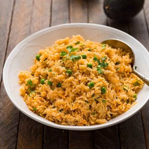 A bowl of arroz roja on a dark wood background. The red rice is garnished with cilantro and green peas.