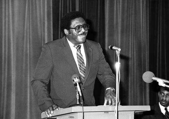 A black and white photo of a smiling Mr. Clark wearing a striped suit as he stands at a lectern before microphones.