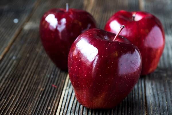 Tři lesklá červená jablka Red Delicious na rustikálním dřevěném povrchu, jedno jablko v popředí a dvě mírně rozmazaná v pozadí.