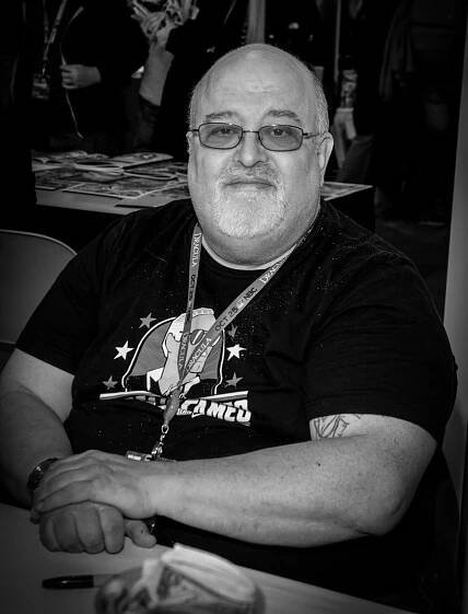 A black-and-white photo of Peter David, a heavyset bald man with a white goatee and tinted glasses. He is seated at a table and wearing a black T-shirt with a design on it.