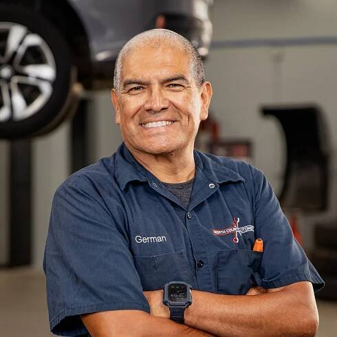 A man in a blue shirt is standing in front of a car with his arms crossed.