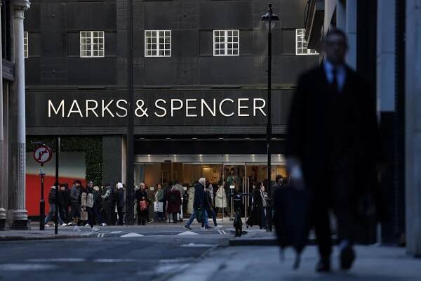 A person wearing a suit and carrying a briefcase walking in the foreground while in the background a group of people stand outside a building with “Marks & Spencer” on it.
