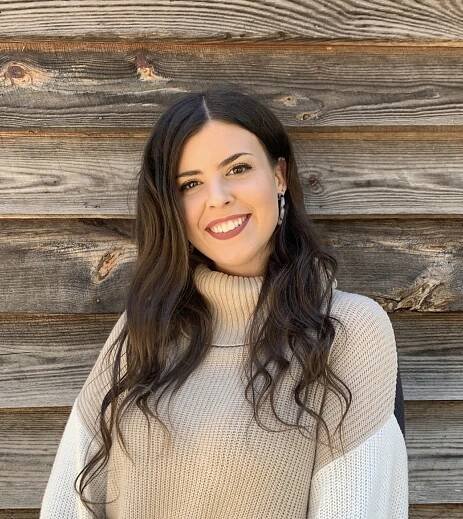 A woman in front of a wooden building wearing earrings and a beige and white turtleneck sweater.