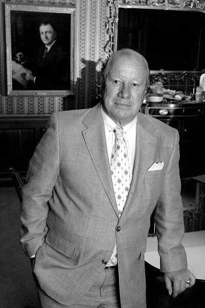A black-and-white photo of Mr. White, a balding man with white hair, standing in front of a wall with a portrait of a man and a mirror on it. He wears a suit and tie.