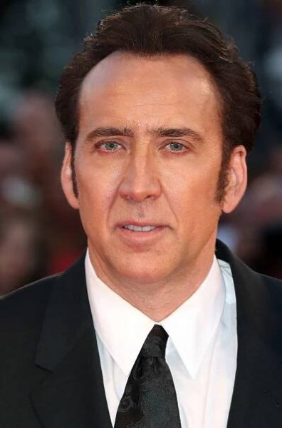 Nicolas Cage with long side burns while wearing a black suit and tie with a white button-down.