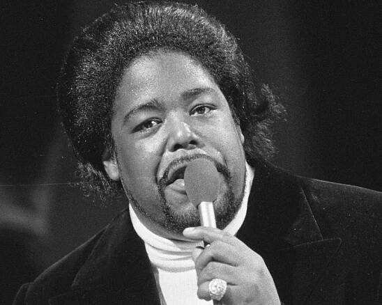 Barry White holding a microphone with a big ring on his pinky, in a turtleneck sweater and blazer, in a black-and-white photo.