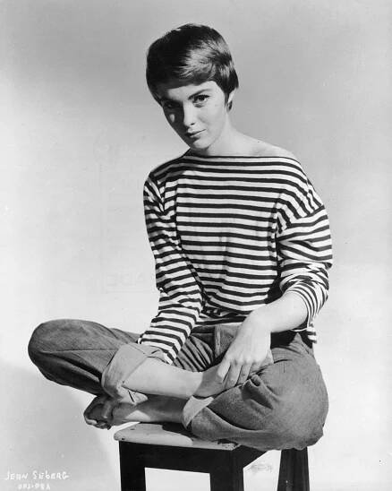 circa 1965: Promotional portrait of American actor Jean Seberg (1938 - 1979) sitting barefoot and cross-legged on a stool, wearing rolled blue jeans and a French-striped sailor jersey pulled off one shoulder.