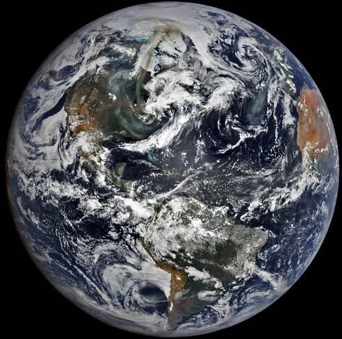Image of the Earth zoomed out to span the entire Americas and part of Africa. The visible land is primarily green to dusky brown in color, aside from Northern Africa, which is a bright tan color. Clouds are interspersed across the entire image obscuring much of the land and blue ocean below, but a slight discoloration between Africa and South America is labeled as dust. A significant darkened swirl coupled against the swirling clouds over North America. This is labeled as smoke.