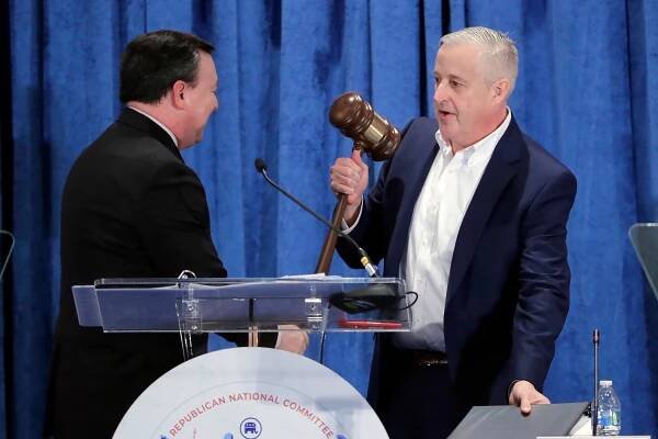 Michael Whatley, right, stands holding a large gavel and talks with a man in a dark suit on a stage. The curtain behind them is blue.