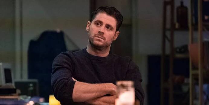 Jon Abrahams looks pensive while sitting at a table with his arms crossed